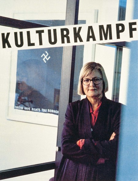 Jenny Harper, as pictured in the New Zealand Listener (5 Sep 1998; v.165 n.3043:p.38-40) with Peter Robinson’s Pakeha have rights too! (1996). This work hung in Jenny’s office at Victoria University, where she staunchly defended its controversial iconography in face of robust criticism from several students and staff who disapproved of the work’s prominent swastika and seemingly racist sentiments. (See Tessa Laird: http://www.physicsroom.org.nz/archive/2cents/hasty.htm)