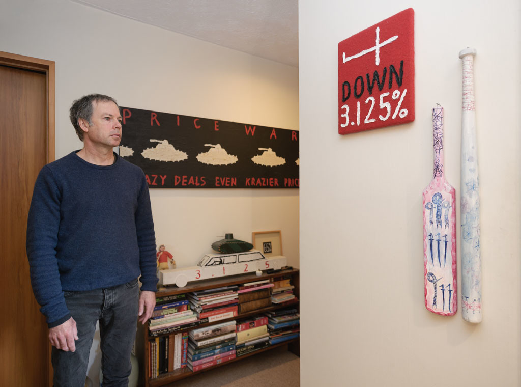 Tony de Lautour with pieces from his art collection. Left hand wall, top to bottom: Peter Robinson Price War 1995. Oil on canvas; Unknown toy tank c. 1950s. Wood and enamel paint; Peter Robinson 3.125% Car. Oil & acrylic on wood; Peter Robinson 65% 1997. Acrylic on wood. Right hand wall, left to right: Peter Robinson Down 3.125% (North & South Edition) 1994. Oil on wool; Tony de Lautour Keeping Score 1997. Acrylic on cricket bat; Tony de Lautour State Security 1997. Acrylic on baseball bat. Photo: John Collie  